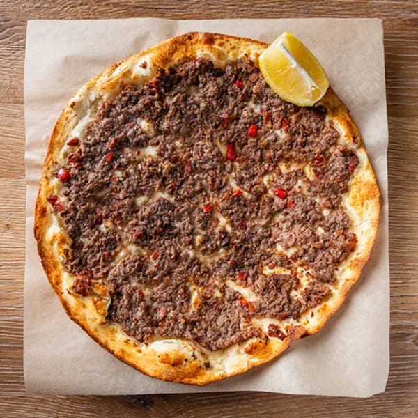 A Lebanese Lahmacun, a round flatbread topped with minced meat and red peppers, with a lemon wedge.