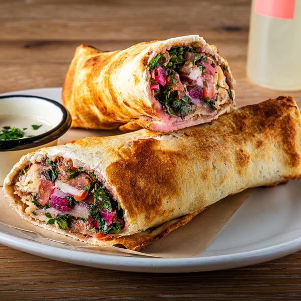 Two Zazzy Lebanese shawarmas, one cut in half, revealing meat, herbs, and pickled vegetables, with a side of sauce.