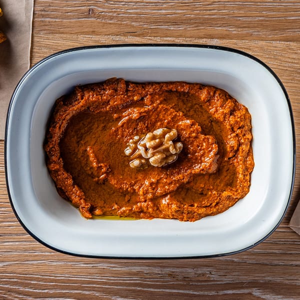 A white rectangular dish of Zazzy's Lebanese muhammara dip, garnished with a walnut, on a wooden table.